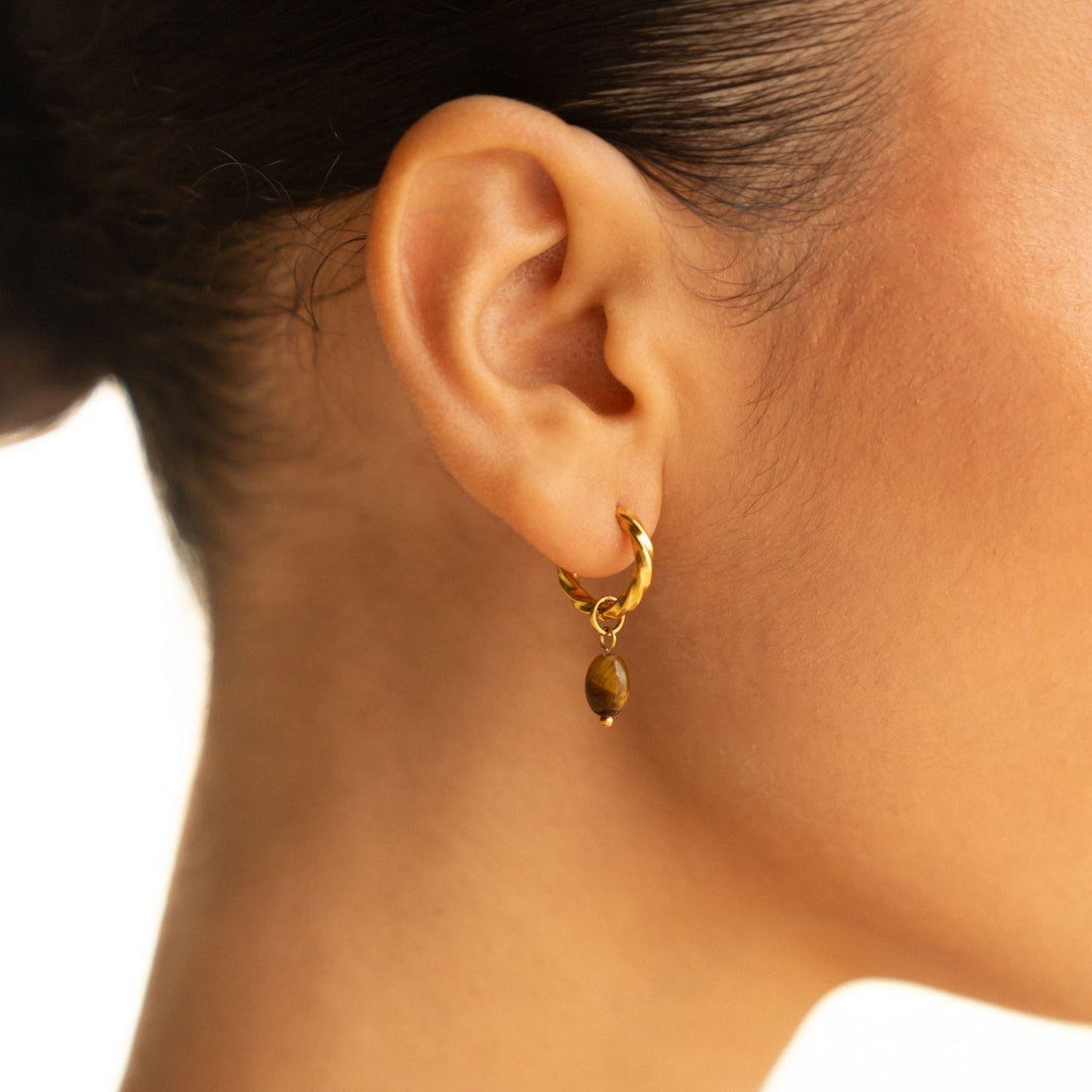 Detailaufnahme von goldenen Huggie Creolen für Damen mit glänzendem Tigerauge Anhänger. Hochwertiger Schmuck.